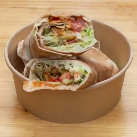Two halves of a falafel wrap stacked in a kraft bowl, cut to show the falafel, salad and tahini inside.