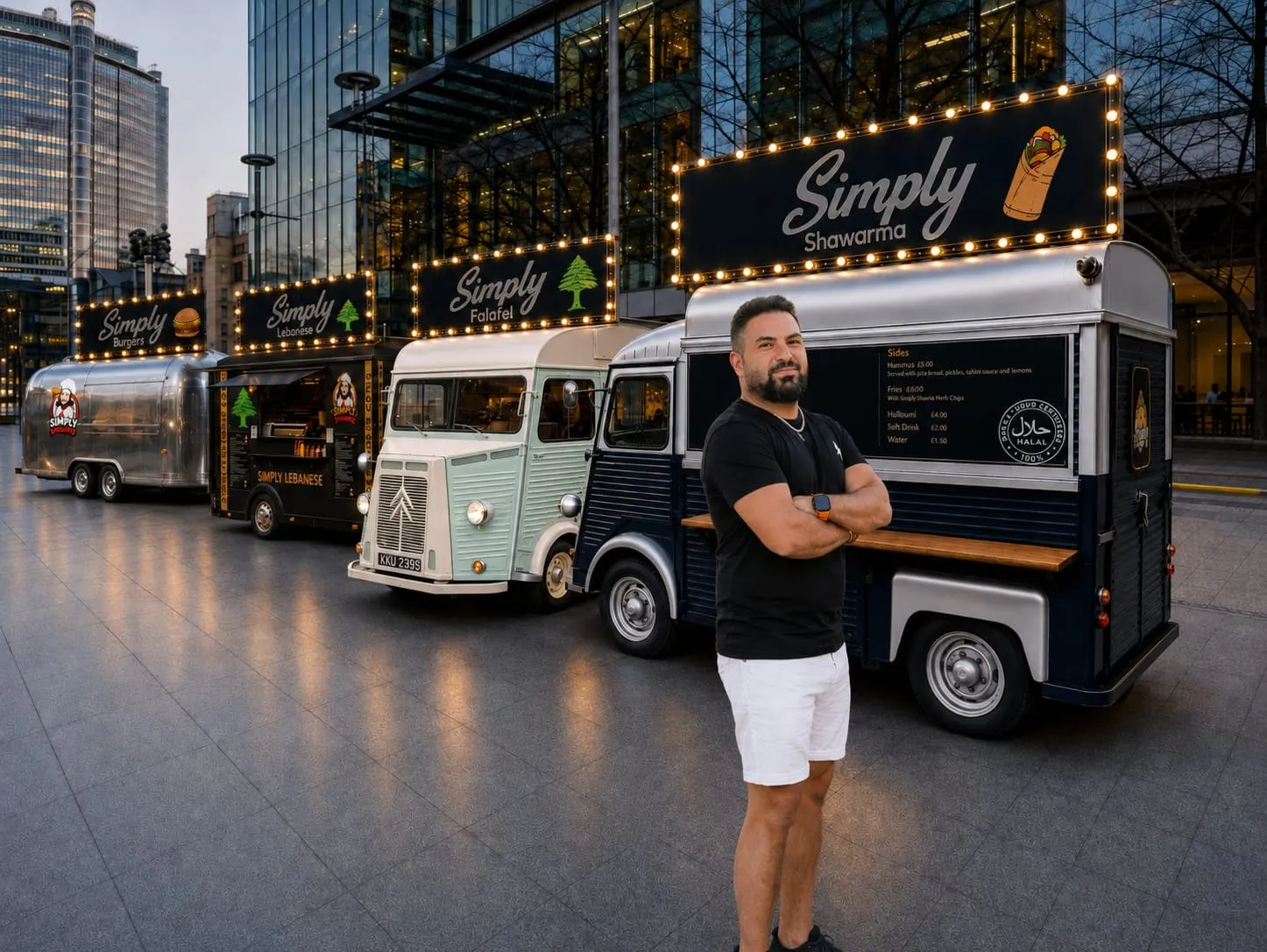 The owner of Simply standing with arms folded in front of the four Simply trucks — Shawarma, Falafel, Lebanese and Burgers — their signs lit up at dusk beside glass office buildings